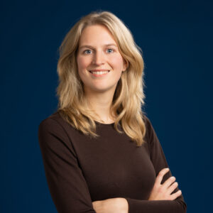 Een vrouw met lang blond haar, gekleed in een donker shirt met lange mouwen, staat tegen een effen donkerblauwe achtergrond en glimlacht met haar armen over elkaar.
