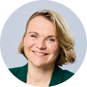 Een glimlachende vrouw met kort blond haar, gekleed in een groene blazer, die naar de camera kijkt tegen een effen lichtgrijze achtergrond.