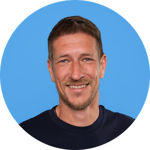 Een glimlachende man met kort bruin haar, een kortgeknipte baard en een snor, gekleed in een donker shirt, die poseert voor een effen blauwe achtergrond.