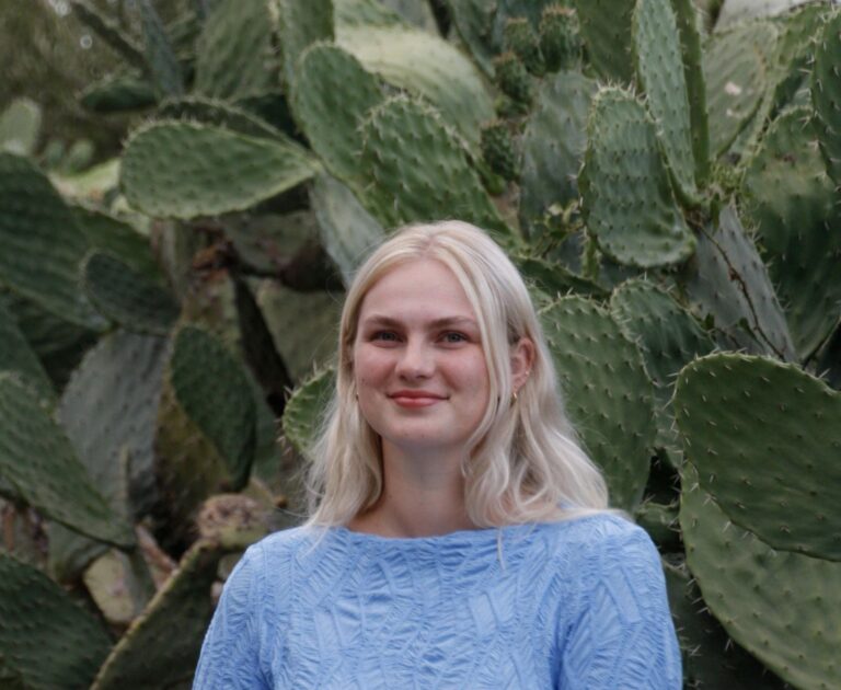 Eine Frau mit blonden Haaren, die ein hellblaues Oberteil trägt, steht vor einem großen Kaktus und lächelt in die Kamera.