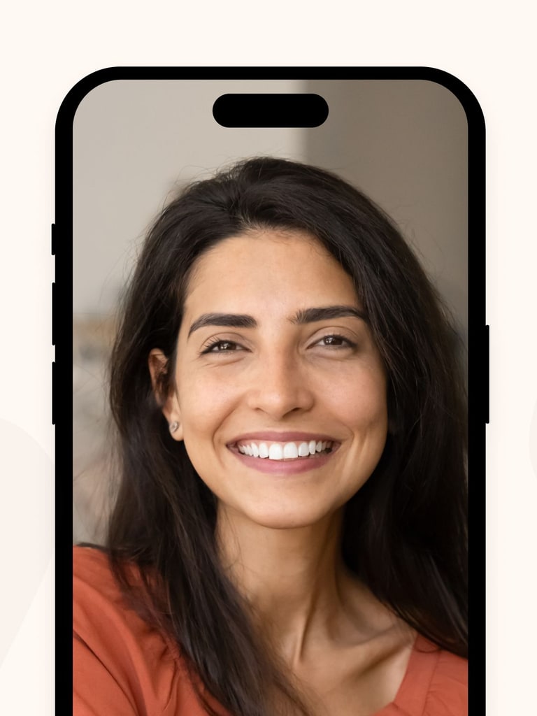 A woman with long dark hair and a warm smile appears on a smartphone screen during a video call, wearing an orange top. Video call icons are visible at the bottom of the screen.